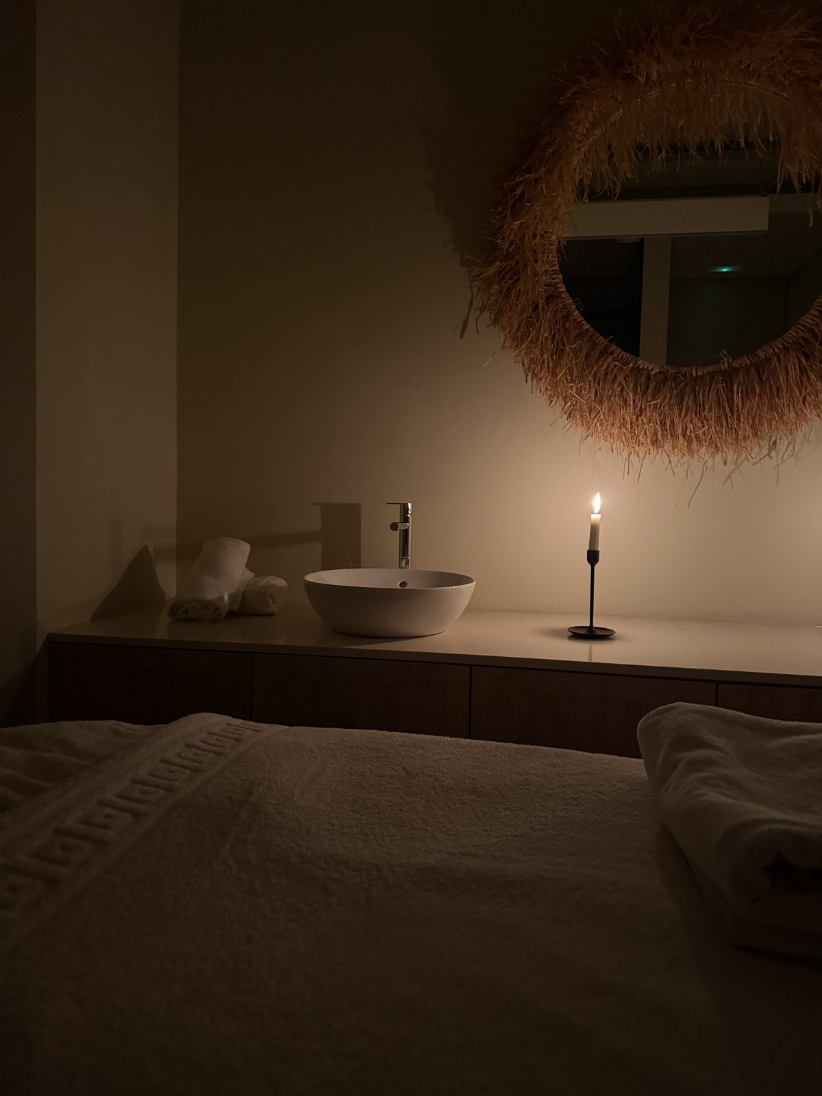A candlelit wellness room with soft towels and a natural woven mirror